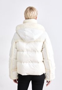 Puffi valkoinen takki, jossa on fleece-vuorattu huppu ja erottuvat tekstuuriset sivupaneelit. Varustettu vetoketjullisilla avauksilla ja pehmeällä pinnalla.