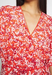 Robe portefeuille florale en orange vif, avec un décolleté en V et des manches courtes bouffantes. Le tissu présente un mélange de motifs floraux rouges et blancs.