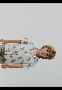 Child with light brown hair wearing a white short-sleeve shirt with orange cherry patterns and beige pants against a plain background.