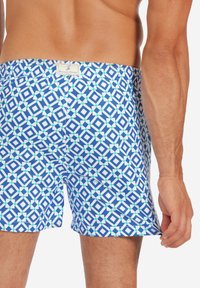 Blaue Badehosen mit geometrischen weißen und grünen Mustern, elastischem Bund und einem kleinen Logolabel auf der Rückseite. Leichter Stoff.