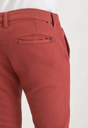 Close-up van een persoon in roestkleurige broek met een klein zwart logo op de achterzak en een witte blouse die erin is gestopt.