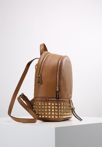 Ruskea nahkareppu, jossa on kultaisia niittejä pohjassa, kaksozip-automaattiset osastot ja säädettävät olkahihnat valkoisella jalustalla.