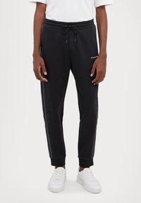 Black sweatpants with a drawstring waist, side white stripes, ribbed cuffs, and embroidered logo on the left. Worn with white sneakers.