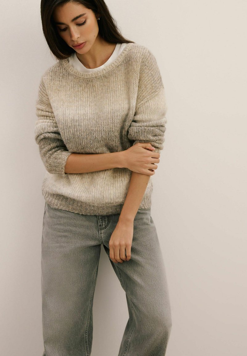 Przesadnie duży beżowy sweter z dzianiny o ribbowanej fakturze, nałożony na biały T-shirt. Połączony z jasnoszarymi, spodniami z wysokim stanem.