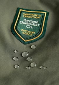 Kaki zunanja jakna iz blaga z zelenim in zlatim vezenim znakem "Portland Outerwear Co." in kapljicami vode na površini.