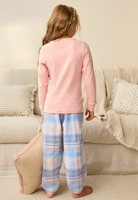 Lyserød langærmet top og blå ternede pyjamabukser med lyserøde og hvide accenter, båret af et barn, barfodet på et lyst tæppe.