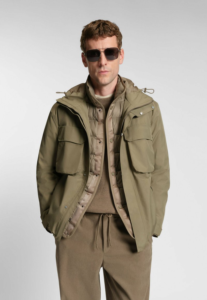 Giacca verde oliva con cappuccio, doppiopetto, con due grandi tasche frontali, indossata sopra un maglione beige, abbinata a pantaloni coordinati.