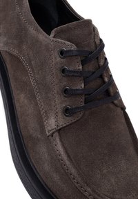 Chaussure en daim marron avec lacets noirs, bout rond et semelle en caoutchouc noir contrastante. Présente une couture sur le côté et des œillets en métal.