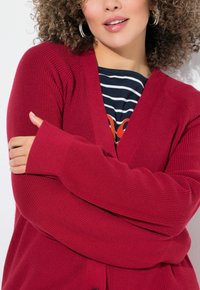 Roter gerippter Cardigan mit V-Ausschnitt, langen Ärmeln und Knopfverschluss auf der Vorderseite. Kombiniert mit einem gestreiften Oberteil darunter und mit weicher Textur.