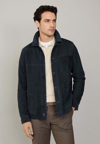 Marineblauwe suedejas met een kraag, knoopsluiting, twee voorzakken en contrasterende stiksels. Gedragen over een beige poloshirt.