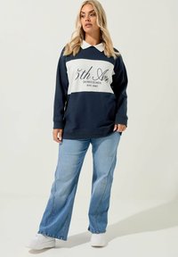 Marinblå och vit långärmad topp med krage, med texten "5th Ave DOWNTOWN EST. 1987", ihop med ljusblå utsvängda jeans och vita sneakers.