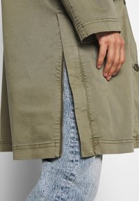 Khaki trencz z bocznym zamkiem i szwami, w połączeniu z jasnoniebieskim fakturowanym dżinsem. Przyszywane rękawy, brak widocznych guzików.