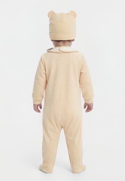 Toddler standing, facing away, wearing a beige footed onesie and matching hat with bear ears against a plain white background.