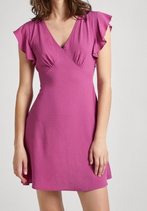 Vrouw draagt een korte roze jurk met fladdermouwen en een V-halslijn, staand tegen een effen lichte achtergrond.