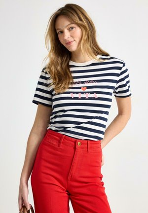 Kvinde med langt lysebrunt hår iført en sort-hvid stribet skjorte med teksten "La dolce VITA" og højtaljede røde bukser, der holder en brun taske.