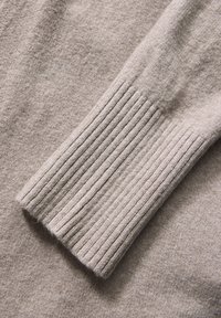 Détail d'un pull en tricot beige avec des poignets côtelés et une texture douce, un corps lisse et une coupe classique, idéal pour le superposition.