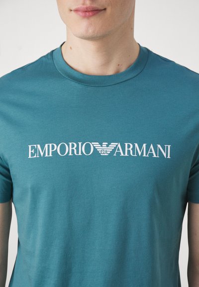 Mėlynos spalvos medvilninė marškinėlių maikė su trumpomis rankovėmis, ant krūtinės puikuojasi balta "EMPORIO ARMANI" logotipas ir grafika.