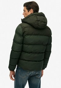 Mørkegrøn puffjakke med hætte, der har vandret syninger og elastiske manchetter. Mærkets logo er placeret på den øverste del af ryggen, og jakken er sat sammen med blå jeans.