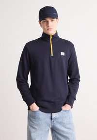 Hanorac navy cu fermoar galben, cu manșete și tiv cu striații, având un mic patch cu logo pe piept. Asortat cu blugi albaștri deschiși.