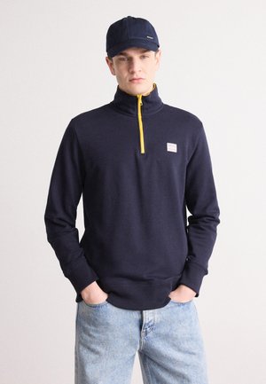 Marinblå ziptröja med gul dragkedja, ribbade muddar och nederkant, med en liten logopatch på bröstet. Matchad med ljusblåjeans.