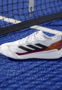 Chaussure de sport blanche Adidas avec des bandes noires et un détail marron au talon sur un court de tennis ou de padel bleu près du filet.