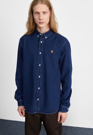 Camisa de mezclilla azul oscura con cuello abotonado, mangas largas y botones blancos. Presenta un pequeño logo naranja en el pecho.