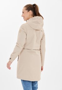 Beige vedenkestävä takki hupulla, joustavat hihansuut, sivutaskut ja takana kiristettävä vyötärömuotoilu. Tasainen rakenne ja huomaamaton brändäysdetalji.