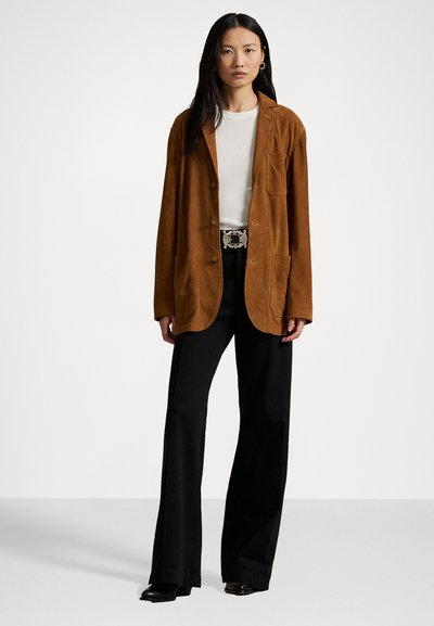Blazer in suede marrone con una vestibilità rilassata, dotato di due tasche frontali, tre bottoni e dettagli di cucitura sottili, indossato su una maglietta bianca.