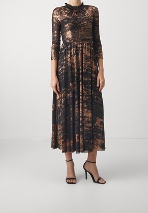 Kobieta ubrana w czarną sukienkę midi z abstrakcyjnym miedzianym wzorem, z marszczoną górą, rękawami do ćwiartki, wysokim kołnierzem i czarnymi sandałami na wysokim obcasie z paskami.