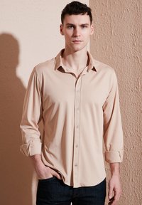 Beige hosszúujjú ing sima anyagból, gombos elülső részsel, felgöngyölt mandzsettákkal és klasszikus gallérral, sötét farmernadrággal viselve.