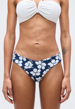 Žena oblečená v bielom bikinovom topu so záhybmi a tmavomodrých bikinových spodkoch s bielym potlačou kvetov ibišteka a tetovaním na ľavom predlaktí.