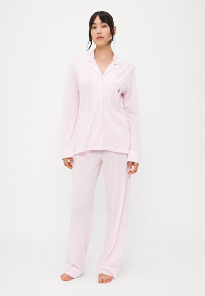 Ροζ σετ πιτζάμας φτιαγμένο από μαλακό ύφασμα· διαθέτει πουκάμισο με κουμπιά και τσέπη, και μακριά μανίκια, που συνδυάζεται με φαρδιά παντελόνια.