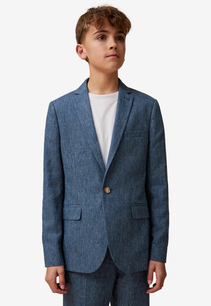Garçon aux cheveux châtain clair portant une veste de costume bleue texturée et un pantalon assorti par-dessus un t-shirt blanc à col rond, regardant légèrement vers le haut.