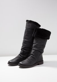 Bottes noires en cuir jusqu'au genou avec un doux revers en fausse fourrure. Design décontracté et semelle plate avec des détails texturés le long du bord.