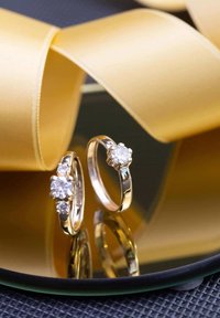 Gold earrings featuring round clear stones, one with a trio of stones in a prong setting, reflecting on a circular black surface.