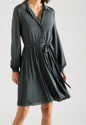 Femme portant une robe plissée manches longues vert foncé avec des boutons et une ceinture nouée à la taille, jupe mi-longue bougeant légèrement.