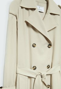 Manteau beige à double boutonnage avec de grands boutons marron foncé, un large col, des épaulettes aux épaules et une ceinture en tissu à la taille.