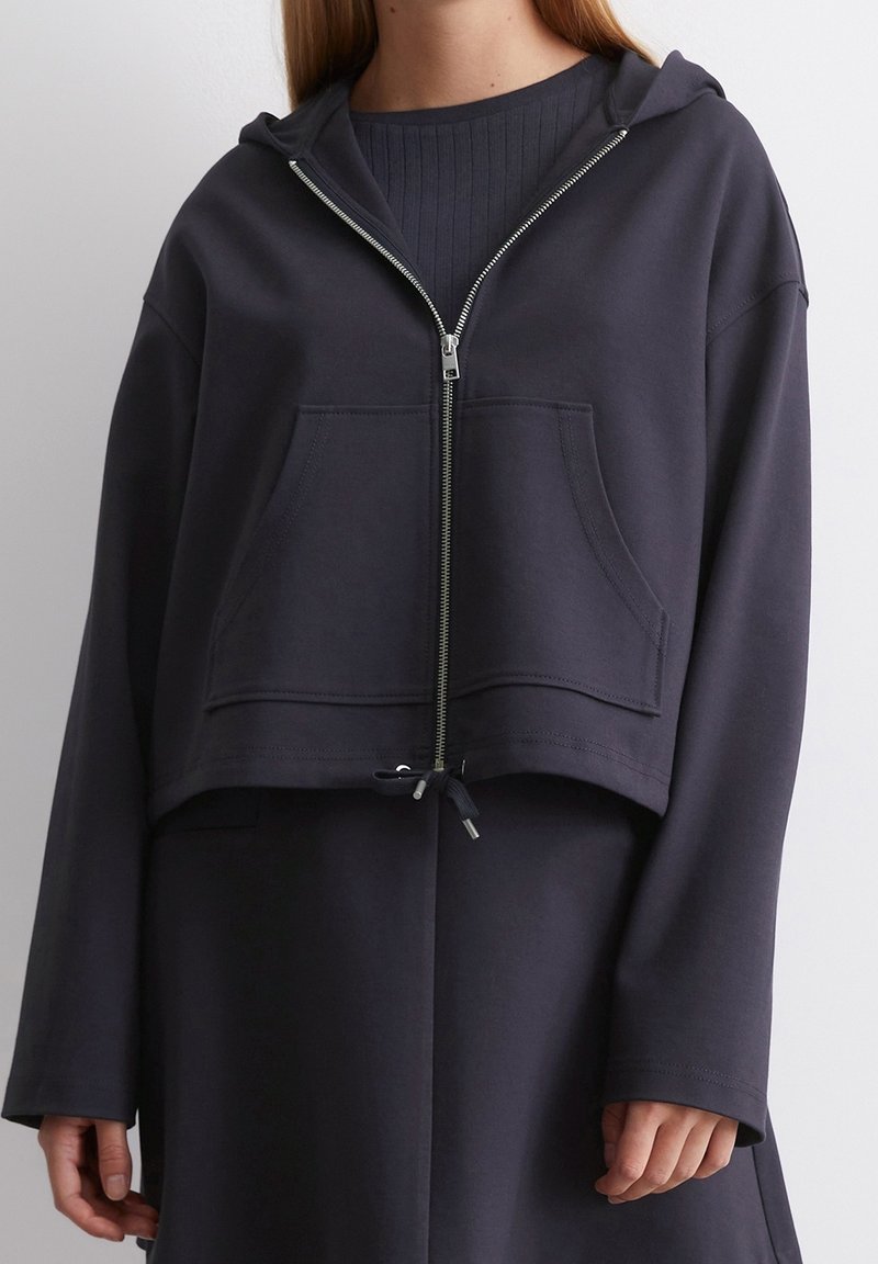 Personne portant un sweat à capuche court bleu marine foncé avec fermeture éclair, poches avant et manches longues amples sur un haut côtelé à col rond.
