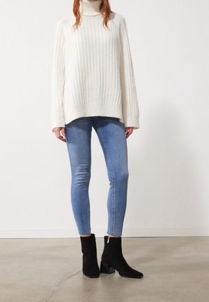 Suéter de cuello alto blanco acanalado con mangas largas, combinado con jeans ajustados azul claro y botines de ante negros con tacones cuadrados.