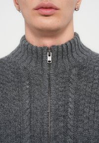 Γκρι πλεκτό πουλόβερ με ψηλό ριμπ κολάρο, που διαθέτει ασημένιο φερμουάρ στο λαιμό. Η υφή είναι χοντρή με υφαντό σχέδιο.