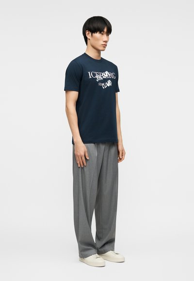 Άνδρας που στέκεται φορώντας μπλε ναυτικό t-shirt με σχέδια, άνετο γκρι παντελόνι και λευκά αθλητικά παπούτσια, μπροστά σε ένα απλό φωτεινό φόντο.