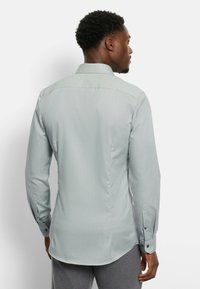 Hellgrünes Langarmshirt mit einem Rückenpasse, geradem Saum und geknöpften Manschetten. Hergestellt aus einem strukturierten Stoff mit vertikalen Streifen.