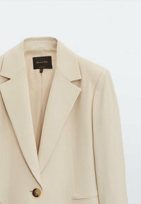 Pehmeä beige bleiseri, jossa on istuva leikkaus, jämäkät kaulukset, yksi nappisulku ja kaksi etutaskua, valmistettu sileästä kankaasta.