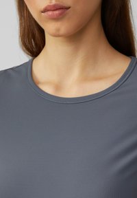 Šedé ribbed tričko s kulatým výstřihem; látka vypadá hladce s jemnou strukturou. Detailní záběr zdůrazňující střih a designové detaily.