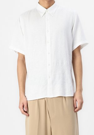 Homme portant une chemise blanche ample à manches courtes boutonnée avec un col et un pantalon beige à jambes larges sur un fond blanc uni.