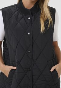 Czarna pikowana kamizelka z stójką, zatrzaskami i bocznymi kieszeniami, prezentująca gładką, matową teksturę.