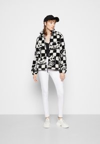 Fato de fleece em padrão xadrez preto e branco com detalhes do logótipo, gola alta, fecho de correr, combinado com jeans brancos e ténis leves.