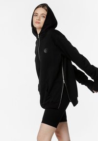 Czarna bluza z zamkiem błyskawicznym i dużą kieszenią z przodu, z teksturowanym logo na klatce piersiowej i dopasowanymi mankietami, zestawiona z czarnymi szortami rowerowymi.