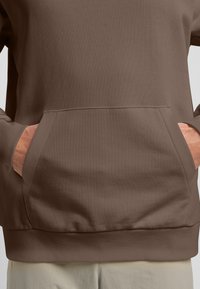 Felpa marrone con superficie tessurizzata, dotata di tasca a marsupio e orlo a coste. Due mani sono posizionate nell'area della tasca.