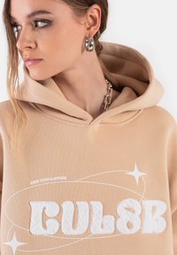 Jasnobeżowa bluza z kapturem z kangurzą kieszenią, z teksturowanym białym napisem i graficznymi akcentami, dopełniona kapturem z regulowanym sznurkiem.